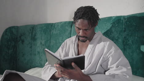 man in white bathrobe sitting on bed reading black notebook, focused on pages, soft light, cozy indoor setting, green velvet headboard