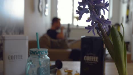 hombre borroso en el fondo sentado en el sofá y tomando café por la mañana con una flor morada enfocada en primer plano