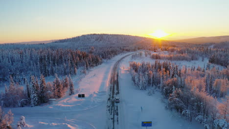 Aerial-view-above-vehicle-joining-snowy-remote-road-heading-towards-woodland-mountain-sunrise
