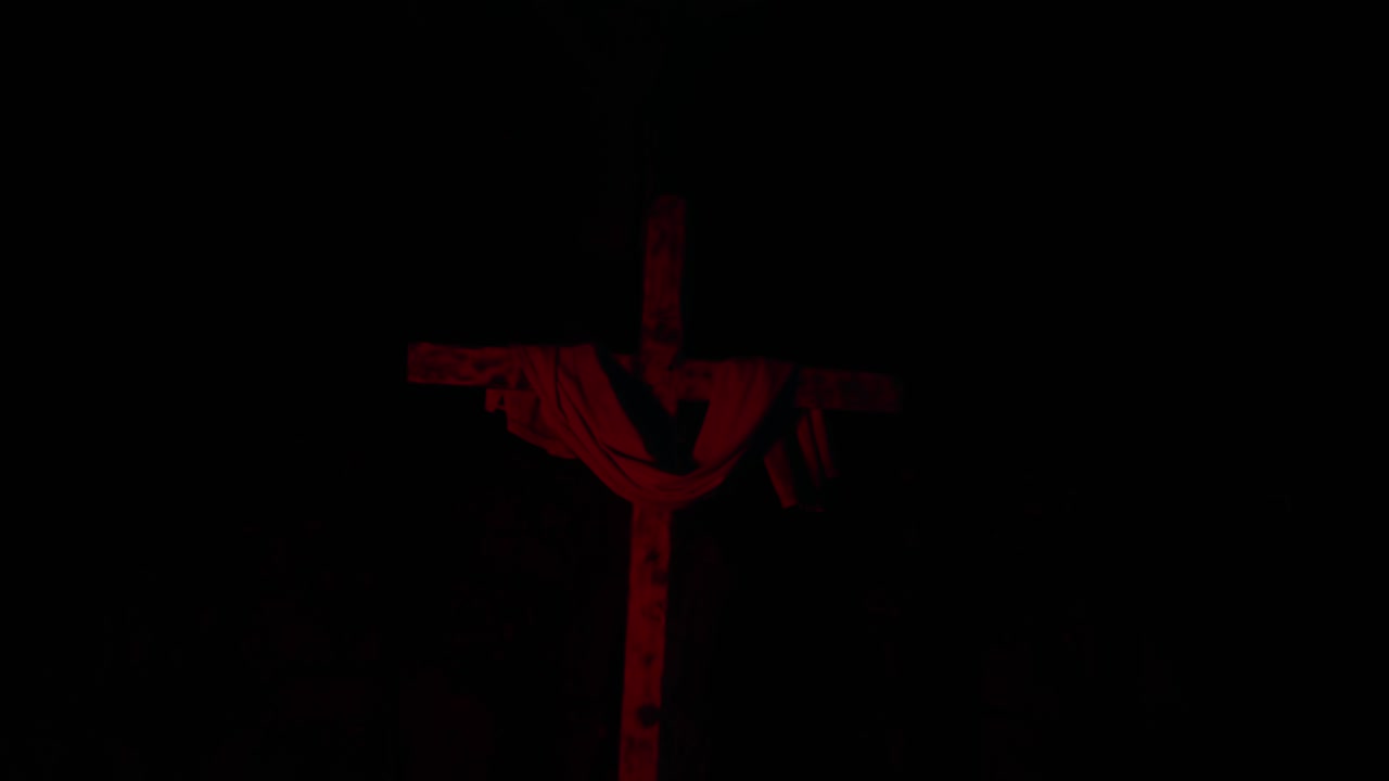 Wooden cross with draped cloth emerging from darkness as deep red light slowly pulses creating a solemn and haunting spiritual atmosphere