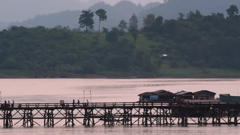 Mon-Bridge-and-a-longboat-followed-speeding-behind-the-structure-while-people-are-walking-on-the-bridge,-silhouetting-as-it-was-getting-dark,-in-slow-motion