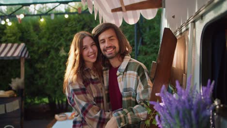 retrato: feliz chico moreno con su novia en una camisa a cuadros abrazándose y hablando mientras está de pie cerca de su remolque durante un picnic en un campamento fuera de la ciudad en verano