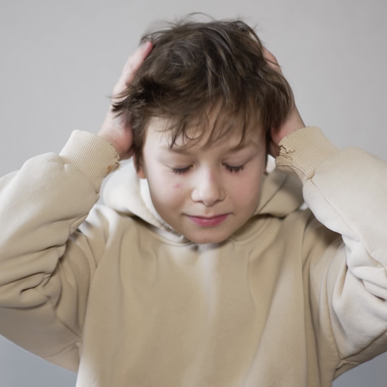 A boy sitting in front of camera in a beige hoodie. Kid takes off his hood and dishevels his brunet hair. Close up