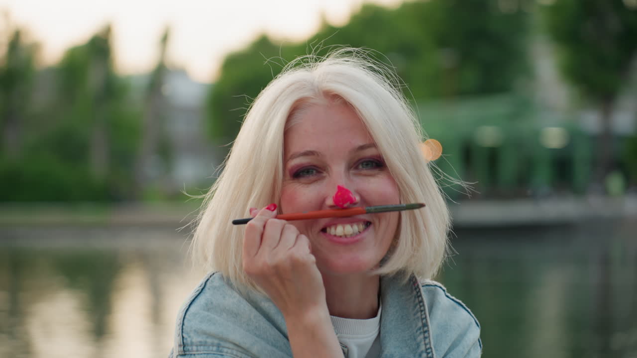 close up portrait of smiling woman balancing paintbrush on painted nose while seated by blurred park waters at golden hour by calm river playful art session captures candid creative bond outdoors
