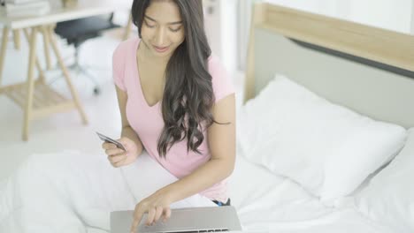 mujer asiática alegre sentada en un cómodo dormitorio, feliz de comprar bienes o servicios en línea, usar tarjeta de crédito haciendo pagos en la tienda de internet en la aplicación en el portátil. concepto de vida cómoda