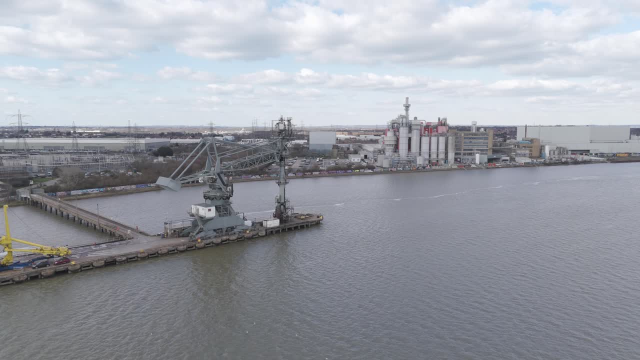 Aerial sweep of riverside chemical factories with cranes, loading arms, smokestacks and storage tanks lining the Thames, illustrating large-scale logistics and processing industry