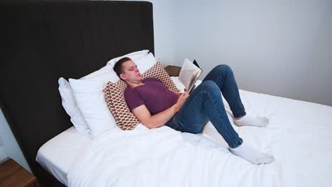 un joven está leyendo un libro con tapa dura negra en su dormitorio, de aspecto serio, acostado en la cama, pasa su tiempo libre con un libro, relajándose en casa, pasa la página, pasa la página