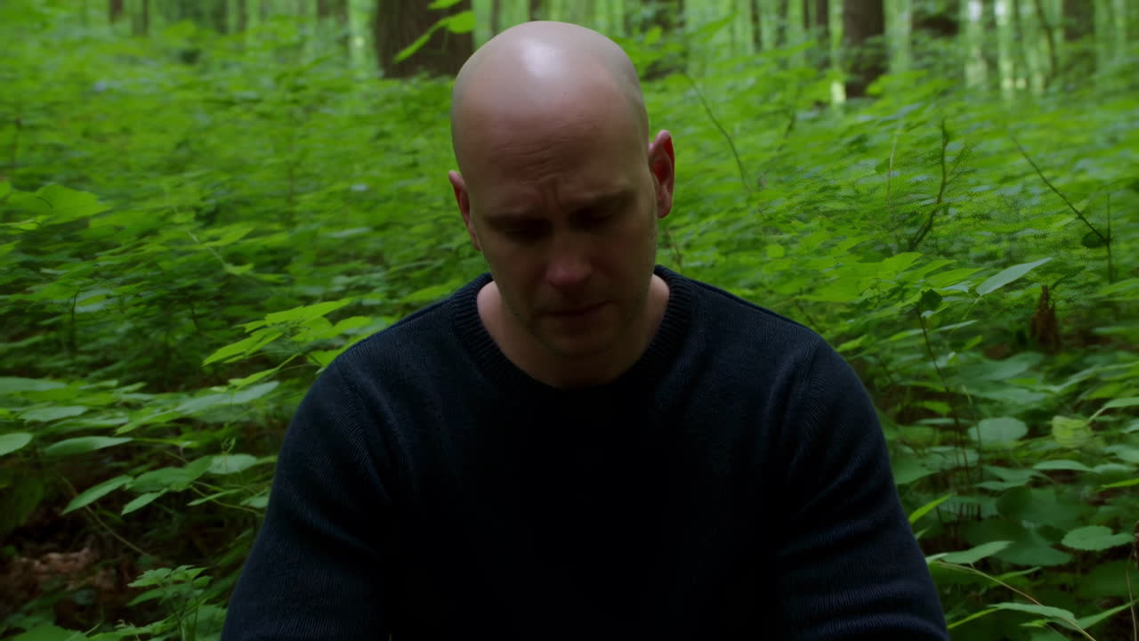 Hvid mand sidder i skov, skaldet voksen og solo vandrer holder pause i tæt grønt, eftertænksomt udtryk.