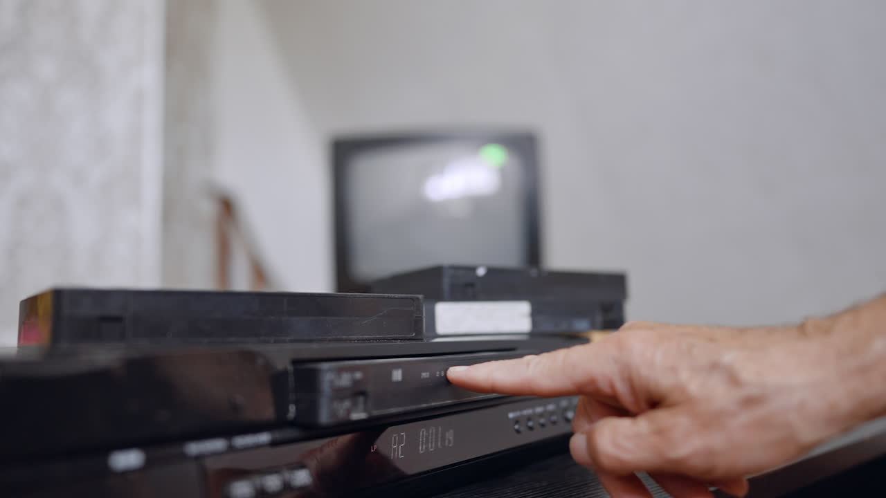 Video Home System with tapes on top. Male hand takes a tape, puts it into the player and pushes the button. Screen at blurred backdrop switches on