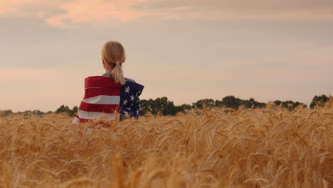 woman with usa flag on her shoulders stands in a wheat field top view