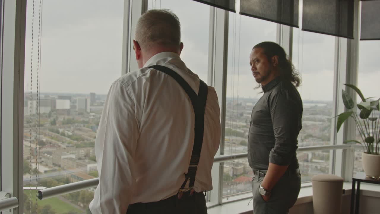Two businessmen stand in front of an office window overlooking a city skyline and talk.