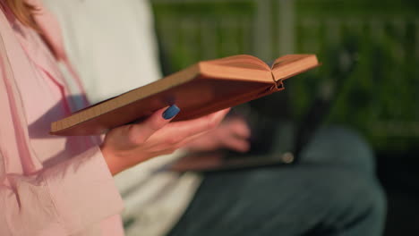 close-up of woman in pink reading from a book outdoors, manicured hands visible, friend in blur typing on laptop, green blurred background enhancing focus