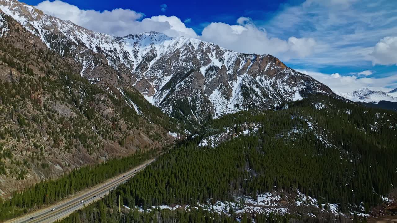 Frisco Gulch ridgeline and winding forest trail with snowy mountains above highway, aerial establishing