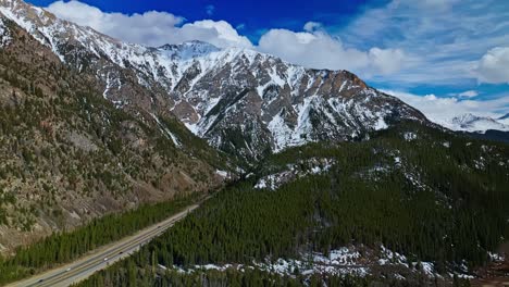frisco gulch ridgeline and winding forest trail with snowy mountains above highway, aerial establishing