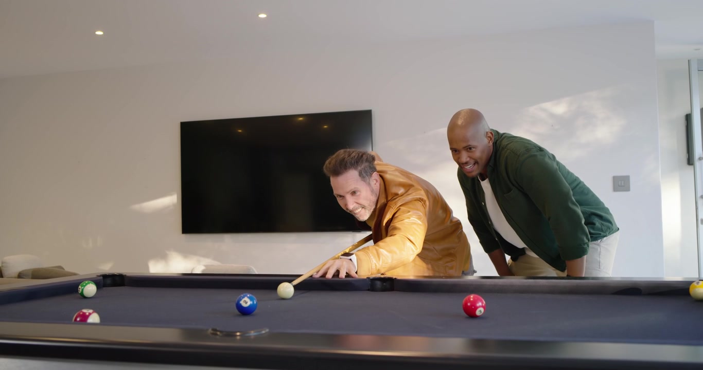 Diverse men joking, shooter leaning over pool table and striking cue ball for fun at home