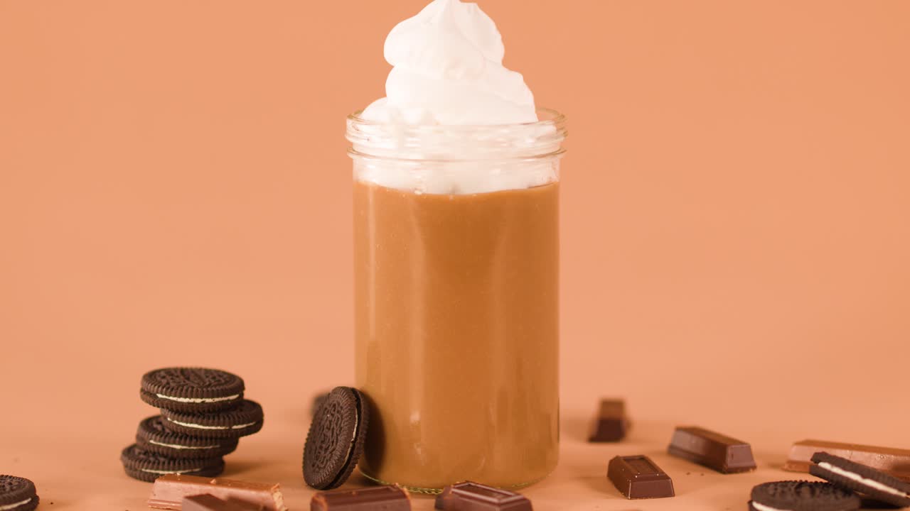 Hand adds chocolate bar to whipped cream milkshake in jar, with cookies and chocolate nearby