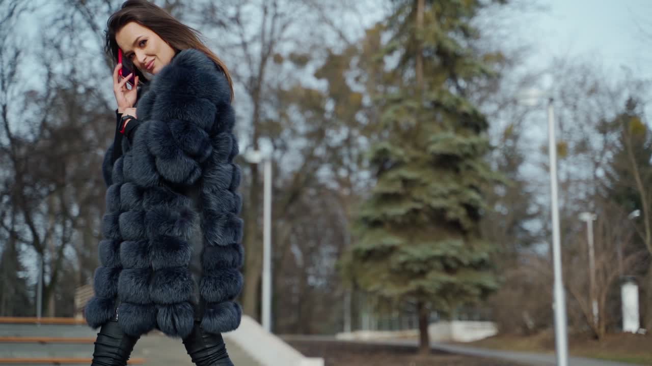 Luxury woman in fur coat. Beautiful young model walking in the park with a phone and looking at camera in cold season. Slow motion.