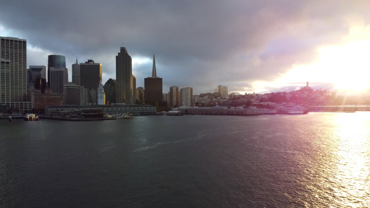 Colorful Sunset Sun Rays Shine Against the San Francisco Bay and Sunlit City Skyline Architecture - Peaceful Pacific Bay Water Waves - Panning Left - Smooth Flying 4K 30fps Aerial Drone Footage