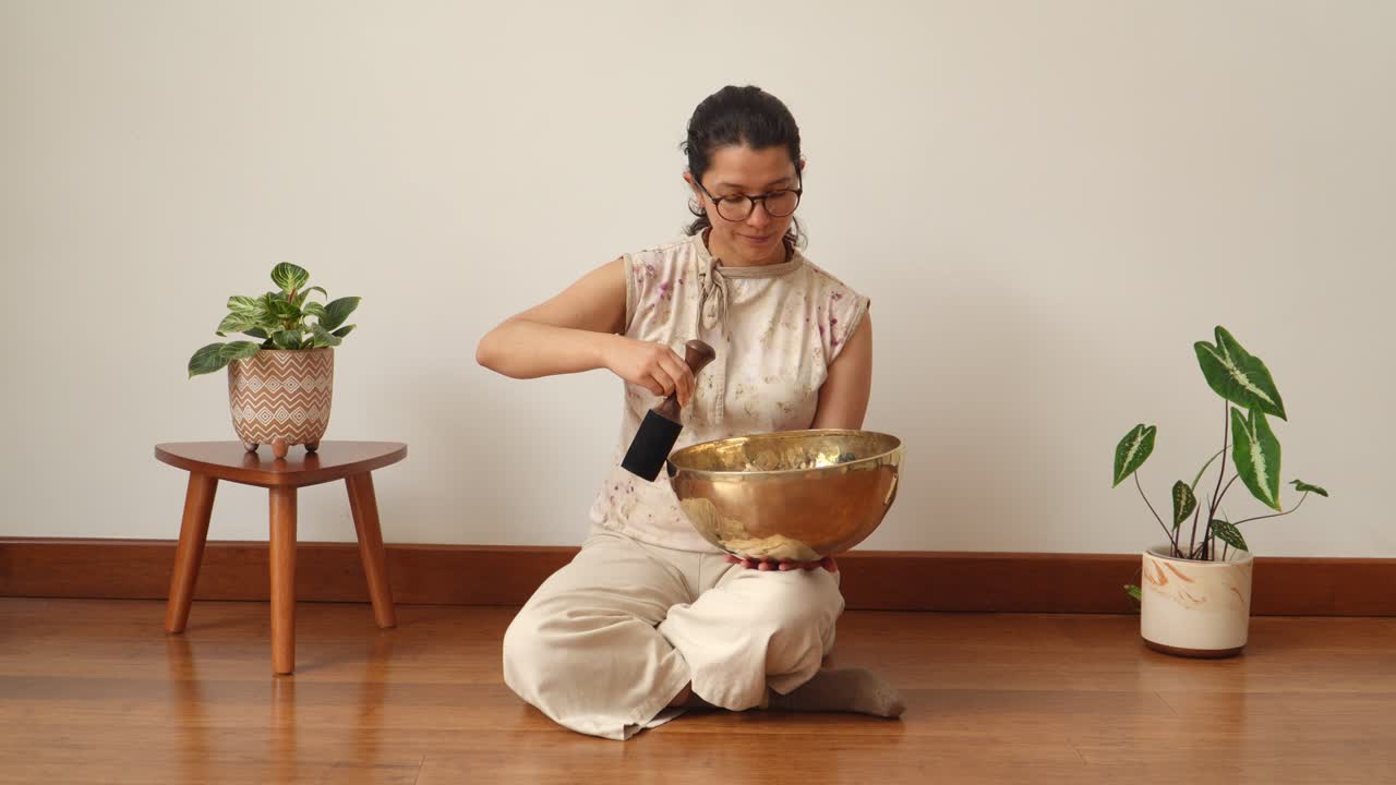 Wide view of a seated woman playing a large Tibetan singing bowl with wooden mallet in a calm minimalist indoor studio