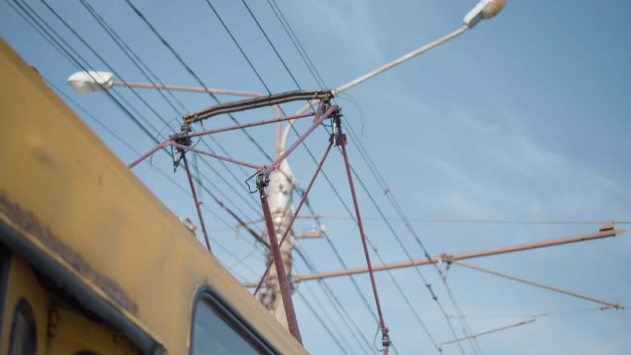 upward view from moving train shows pantograph on overhead wires under blue sky, winter city beyond, snowy street below, yellow roof and poles vibrating as electric transport glides forward