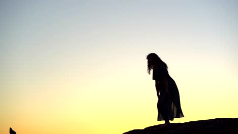 Female-silhouette-walking-on-rocky-hilltop-at-picturesque-sunset