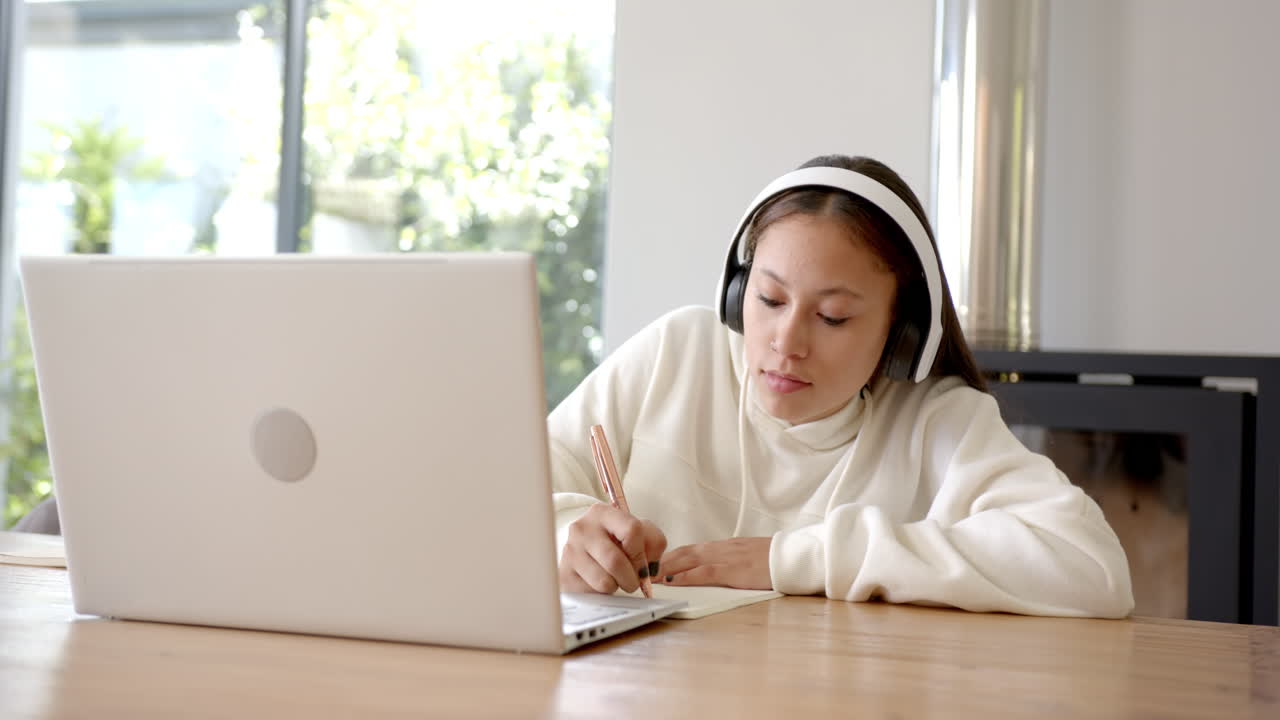 Wearing headphones, girl studying at home with laptop and notebook, copy space