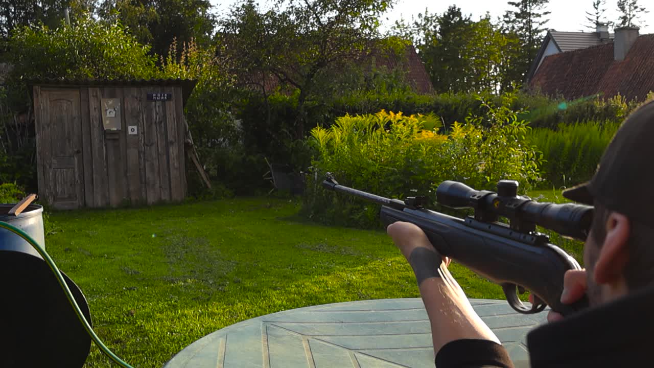 Man with tattoos and black clothing shooting a firearm rifle gun in a sunny sunset green garden in slow motion by aiming through a small scope telescope slowly. lense and light flare visible also