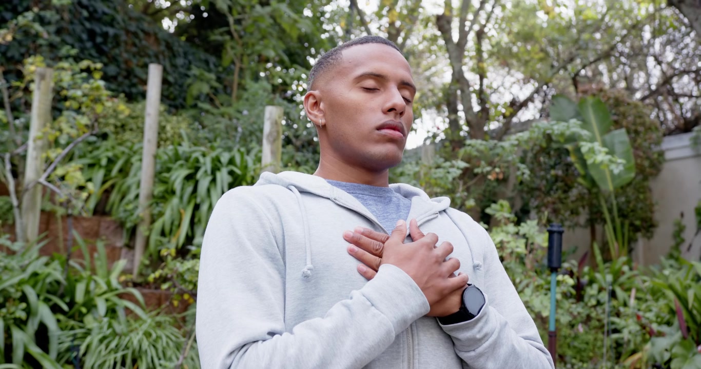 African American man meditating in yard starting hands crossed breathing and tilting wearing hoodie