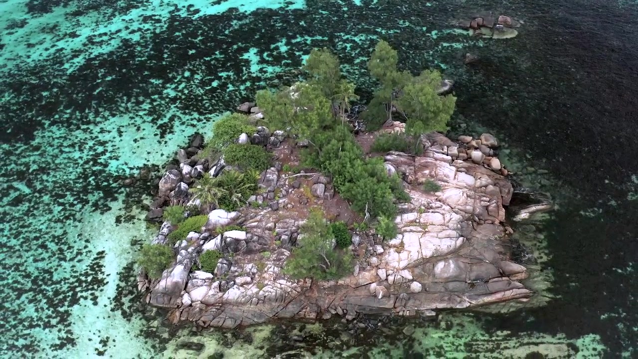 Ripresa aerea a media altezza con drone di un piccolo isolotto roccioso di granito coperto di alberi, interamente circondato da una barriera corallina bassa turchese e da un oceano più profondo nelle Seychelles.