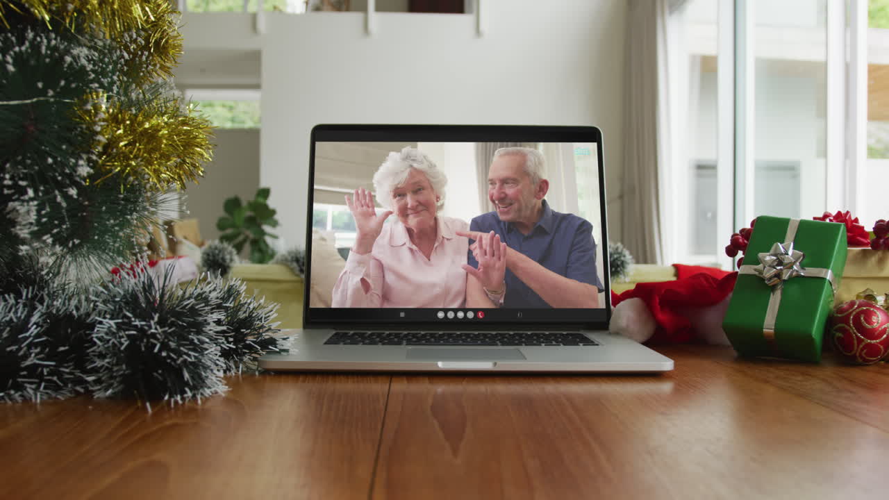 una pareja caucásica de alto nivel sonriente saludando en una videollamada de navidad en una computadora portátil