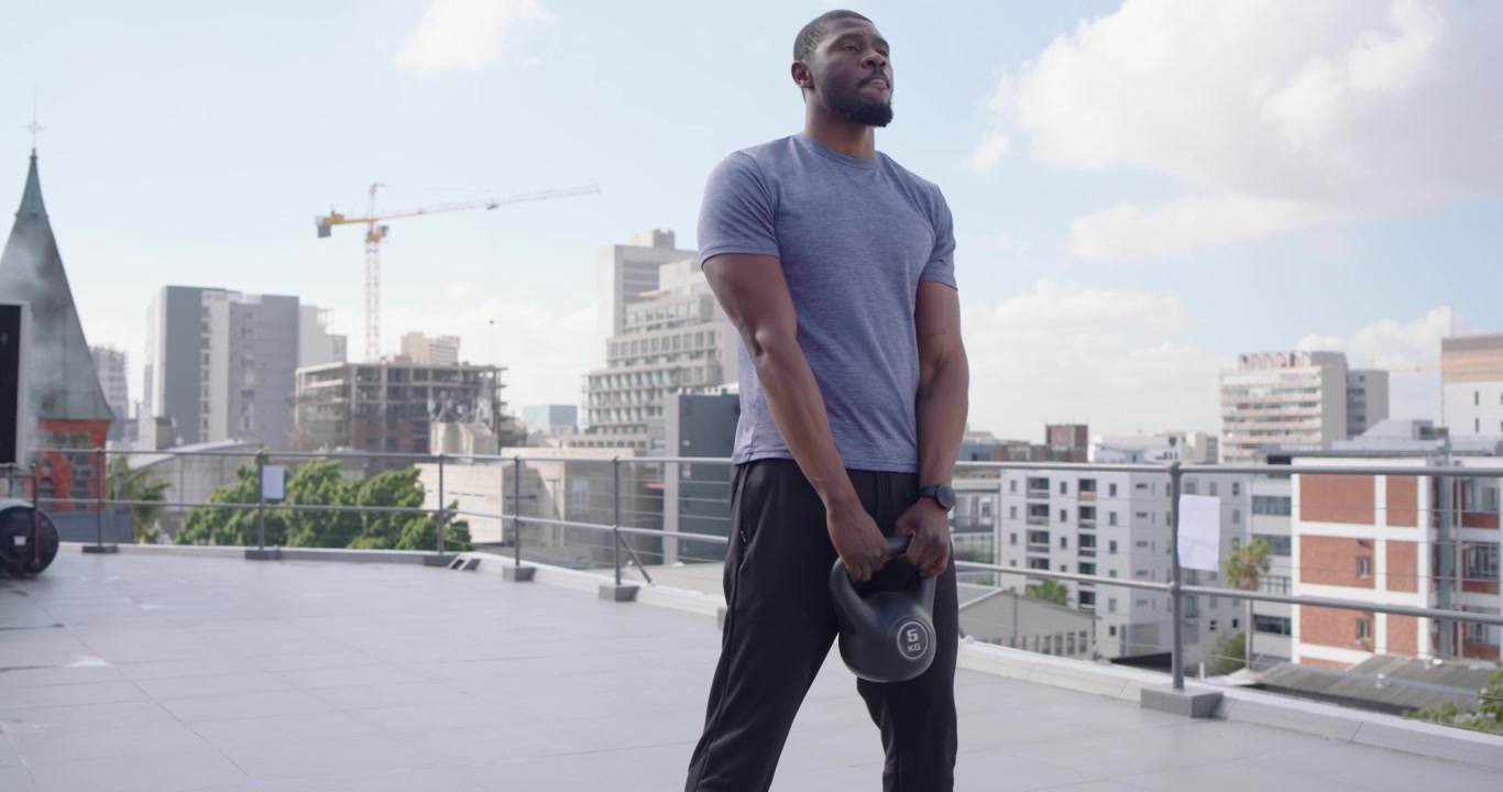 Uomo africano di mezza età che si riscalda con una maglietta blu, poi solleva un kettlebell per il fitness sul tetto.