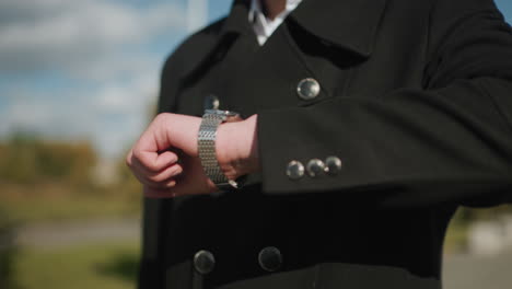 primer plano de una capa negra con botones metálicos brillantes en un entorno urbano al aire libre mientras el individuo comprueba la hora en un reloj de pulsera, con edificios modernos borrosos, coches estacionados y vegetación en el fondo