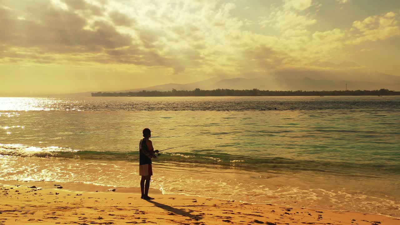 Young man catching fish on sea with fishing rod thrown from tranquil beach at sunset hour on a cloudy sky in Bali coastline