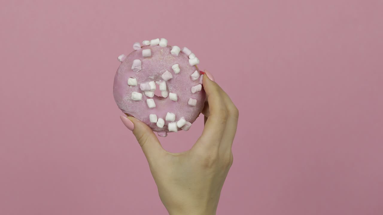 Mano di donna che tiene una ciambella rosa, deliziosa e con zuccherini su sfondo rosa