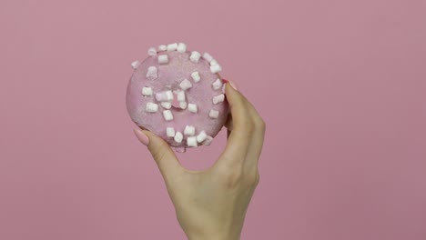 womens hand holding pink, delicious, sprinkled donut on pink background.