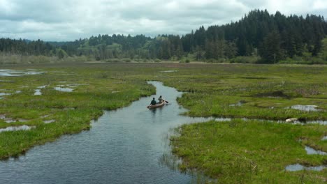 Drone-shot-of-two-people-paddling-a-canoe-through-a-small-waterway-surrounded-by-grass-and-forest