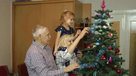 Little-cute-child-girl-with-senior-grandparents-family-decorating-artificial-Christmas-tree-at-home