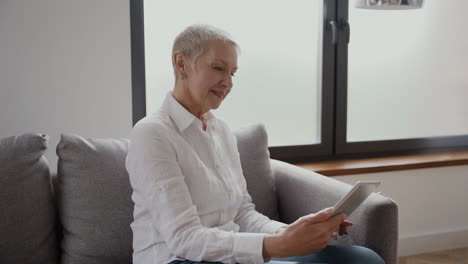 Senior-Woman-With-Short-Blonde-Hair-Sitting-On-The-Sofa-Using-A-Tablet