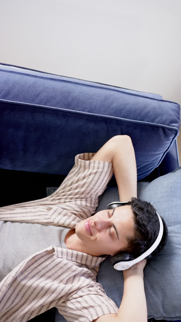 Vertical video of biracial man in headphones lying on couch, copy space, slow motion