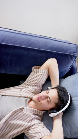 vertical video of biracial man in headphones lying on couch, copy space, slow motion