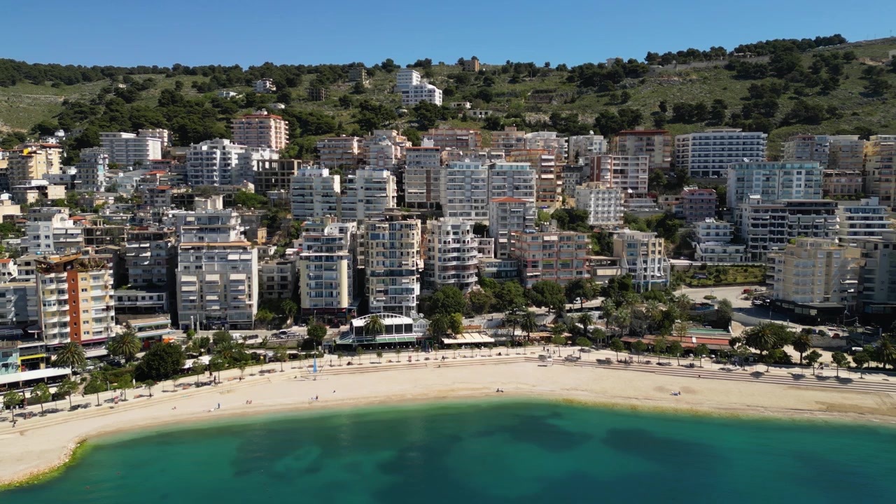 Arnavutluk, Saranda'nın sahil gezinti yolu ve sahil binalarından uzaklaşan havadan drone görüntüleri. Çekim, şehrin modern deniz kenarı mimarisini, kumlu plajı ve turkuaz İyon Denizi'ni ortaya koyuyor.