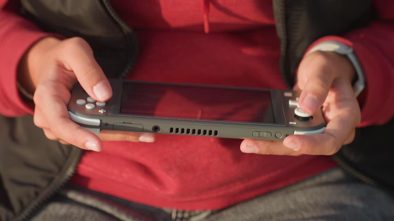 Primo piano delle mani che tengono una console portatile, giovane giocatore con felpa rossa seduto con il dispositivo in grembo, pollici che premono pulsanti e joystick, concentrazione focalizzata e postura rilassata, luce solare calda.