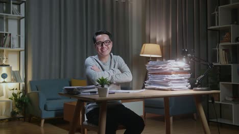 hombre asiático cruzando los brazos y sonriendo a la cámara mientras trabaja con documentos en casa
