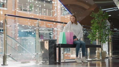 young lady seated in bright modern mall retrieves yellow-covered book from pink shopping bag, surrounded by colorful bags, decorative plants, glass railings, with scattered light reflections