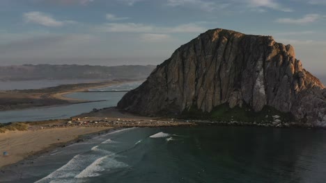 Vista-Aérea-De-Morro-Bay-Rock-En-California-Usa-Durante-La-Puesta-De-Sol-Con-Islas-Arenosas-Y-Montañas-En-La-Distancia