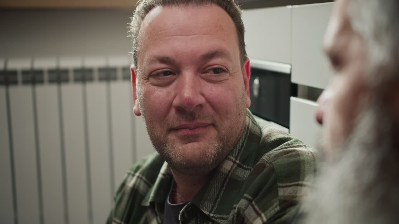 y cerrar un hombre moreno con rastro del universo en una camisa a cuadros se comunica y mira a su novio mayor con una barba gris sentado en el suelo en la cocina. un hombre mayor acaricia la mejilla de su novio con rastro en una pareja lgbt