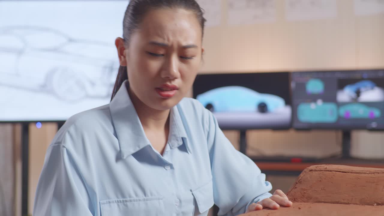아시안 여성 자동차 디자이너가 머리가 아파서 지이를 사용하여 표면을 매끄럽게하고 스튜디오에서 자동차 점토 조각에서 세부 사항을 만니다.