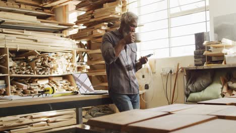 African-american-male-carpenter-using-smartphone-while-drinking-coffee-in-a-carpentry-shop