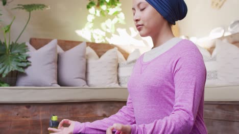 video de una mujer biracial tranquila en hijab sentada en una postura de meditación de yoga con los ojos cerrados en casa.
