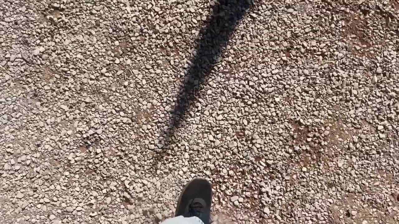 Point-of-view shot of a person stepping over a yellow cable protector on gravel, with black shoe, light jeans, and outdoor event safety detail.